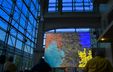 Photograph of a screen displaying a video projection inside the atrium of a glass building. Photograph of a screen displaying a video projection inside the atrium of a glass building.