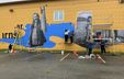 Image of the mural-making process on a yellow wall, featuring a photographic collage of images and typography unified by a flowing graphic form resembling water or a wave. Image of the mural-making process on a yellow wall, featuring a photographic collage of images and typography unified by a flowing graphic form resembling water or a wave.