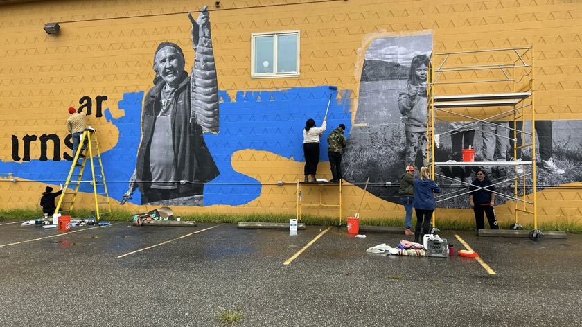 Image of the mural-making process on a yellow wall, featuring a photographic collage of images and typography unified by a flowing graphic form resembling water or a wave.