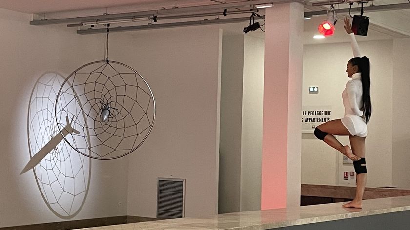 Photograph of a performance showing a dancer in a geometric pose with one leg raised, facing an installation resembling a dreamcatcher with a rocket at its center.