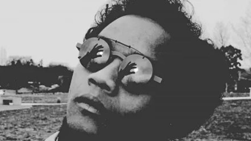 Photograph of a person wearing round glasses, whose lenses reflect their hand holding the sun between their fingers.