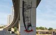 Photograph by Anastasia Spirenko showing a tall highway overpass column extending toward the horizon against an urban landscape. The column features graffiti of an index finger pressing a surface, with red liquid spreading outward from the point of pressure.