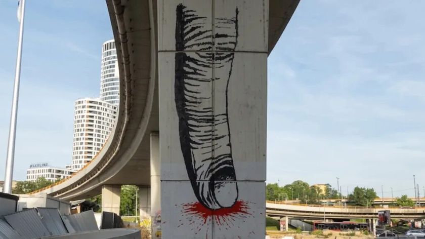 Photograph by Anastasia Spirenko showing a tall highway overpass column extending toward the horizon against an urban landscape. The column features graffiti of an index finger pressing a surface, with red liquid spreading outward from the point of pressure.