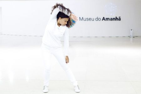 Photograph of a female performer dressed in white against the white interior of a museum. She leans to one side with her right hand behind her head. The words “Museu do Amanhã” are visible on the wall. Photograph of a female performer dressed in white against the white interior of a museum. She leans to one side with her right hand behind her head. The words “Museu do Amanhã” are visible on the wall.