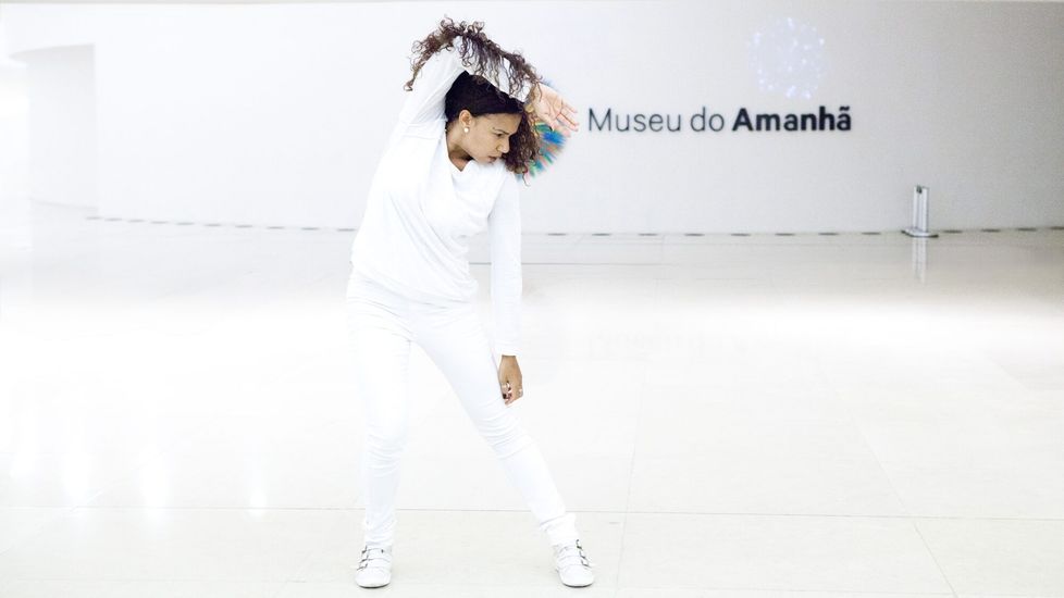Photograph of a female performer dressed in white against the white interior of a museum. She leans to one side with her right hand behind her head. The words “Museu do Amanhã” are visible on the wall.
