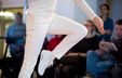 Photograph of a dancer in motion, dressed entirely in white, captured mid-jump with one leg raised in a geometric pose.