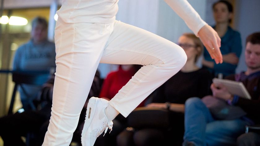 Photograph of a dancer in motion, dressed entirely in white, captured mid-jump with one leg raised in a geometric pose.