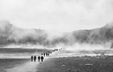 Black-and-white photograph by Sébastien Goldberg showing people walking along a flat road toward mountains on the horizon, surrounded by fog.