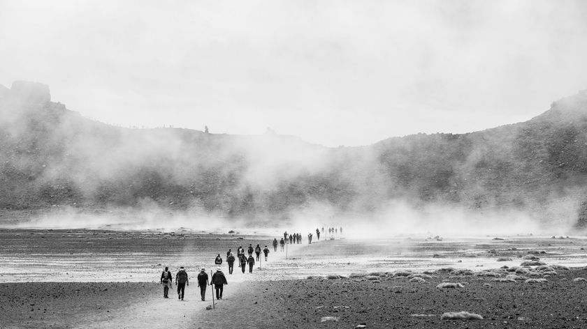 Black-and-white photograph by Sébastien Goldberg showing people walking along a flat road toward mountains on the horizon, surrounded by fog.