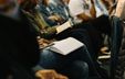 Photograph of people seated in an auditorium with their faces out of view, holding books and notebooks on their laps, evoking an atmosphere of learning.