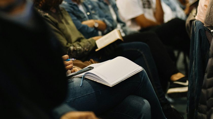 Photograph of people seated in an auditorium with their faces out of view, holding books and notebooks on their laps, evoking an atmosphere of learning.