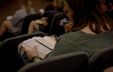 Photograph of a woman sitting in a lecture hall with a book, viewed from behind so her face is not visible.