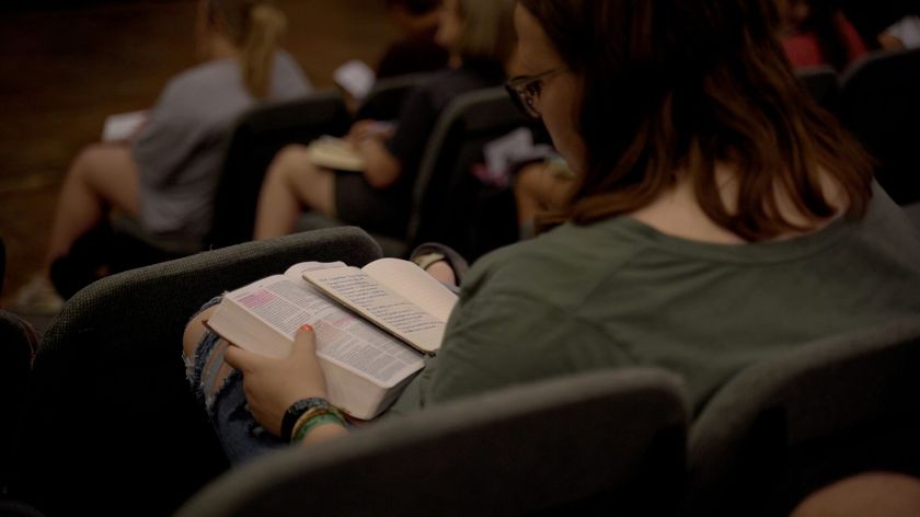 Photograph of a woman sitting in a lecture hall with a book, viewed from behind so her face is not visible.