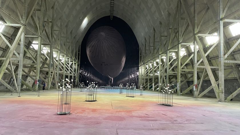 Photograph of a vast interior space containing a large black airship, with an acoustic installation of ten sculptures arranged across the foreground.